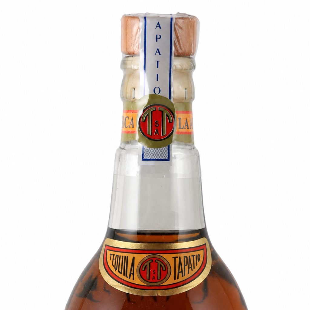 A close-up studio shot of a Tapatio Tequila bottle neck and upper body against a plain white background. The amber liquid, clear glass, cork, and decorative neck labels are clearly visible, with the Tapatio brand name and logo prominently shown.