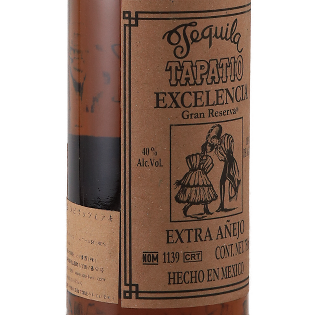 A close-up of a Tapatio Tequila bottle showing the brown kraft paper label with black text and illustration, with the amber liquid visible through the glass against a white background.